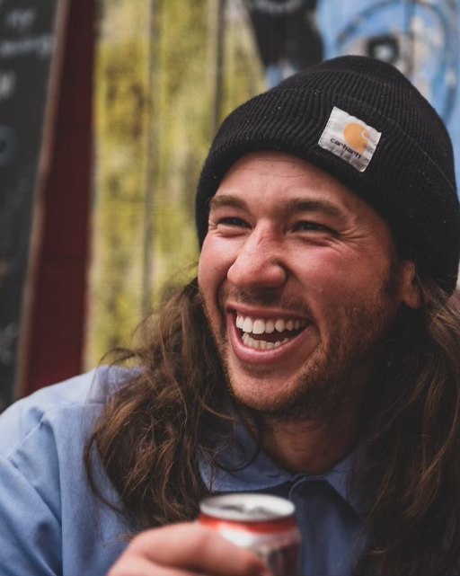 Chris Denesha laughing, in a dark knit beanie, in front of a painted mural wall.
