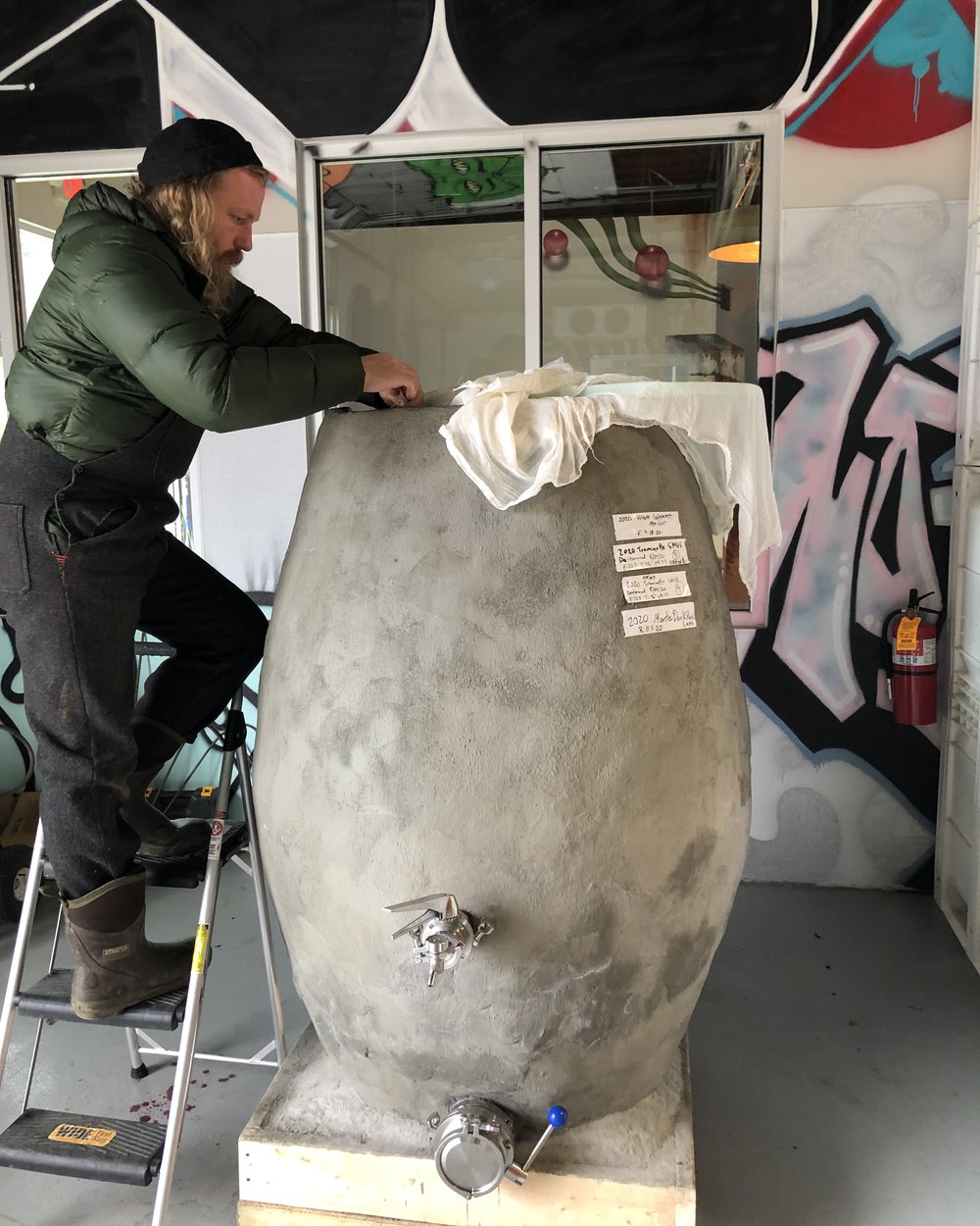 Chris on a stepladder in a winter coat and knit cap, tending a tall concrete egg fermenter draped in cheesecloth, a painted mural wall behind.